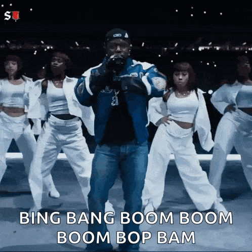 a man in a blue jacket is standing in front of a group of dancers and says bing bang boom boom