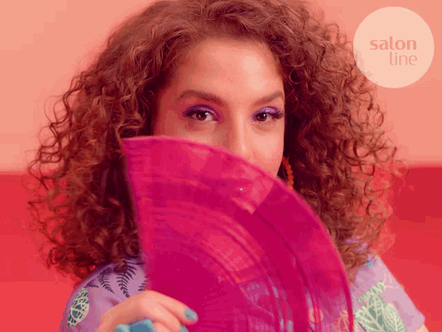 a woman with curly hair holds a pink fan in front of her face with salon line written in the corner