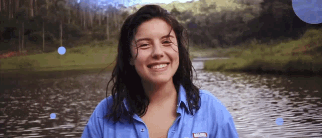 a woman wearing a blue shirt that says brahma smiles in front of a body of water
