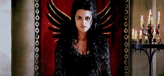 a woman in a black dress is sitting in front of a red wall with a winged banner .