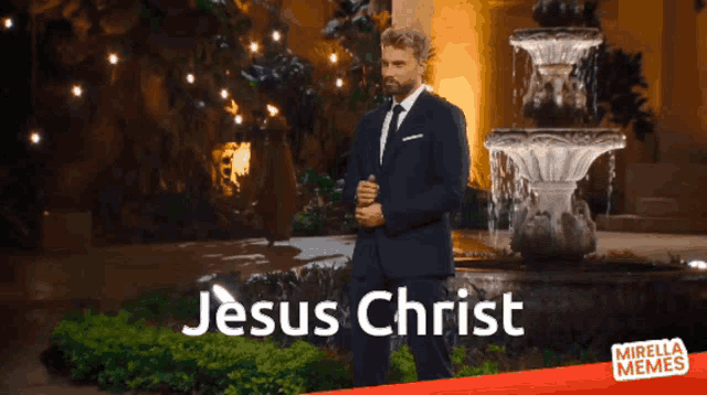 a man in a suit and tie stands in front of a fountain that says jesus christ on it