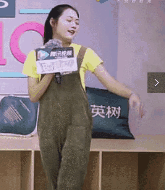 a woman in overalls is holding a microphone in front of a wall with chinese characters on it