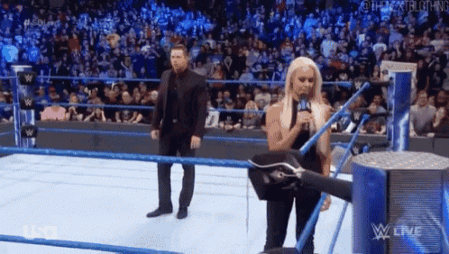 a woman is standing in a wrestling ring talking into a microphone while a man stands behind her