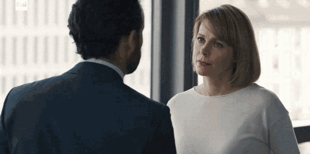 a man in a suit and a woman in a white shirt look at each other in front of a window