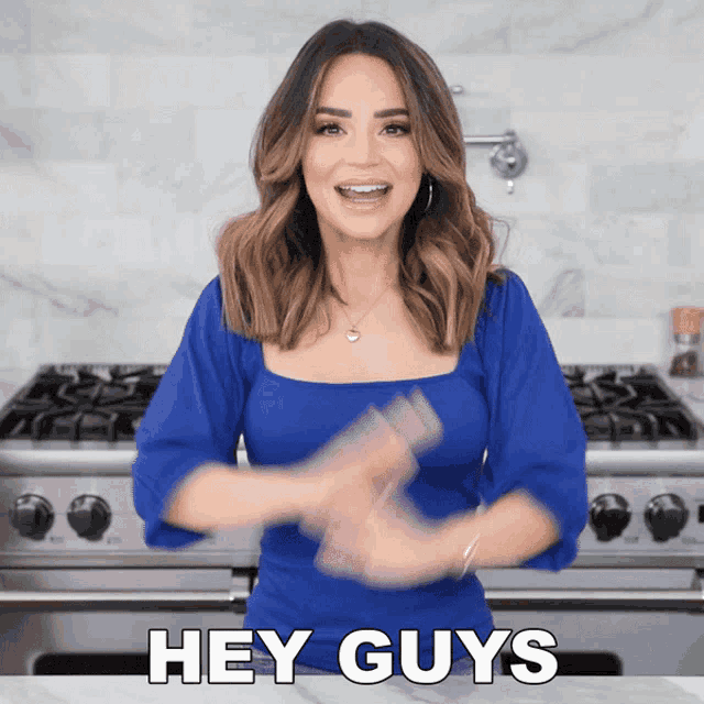 a woman in a blue shirt says " hey guys " in front of a stove