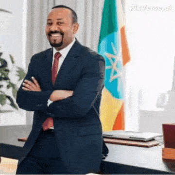 a man in a suit and tie is standing in front of a flag with his arms crossed and smiling .