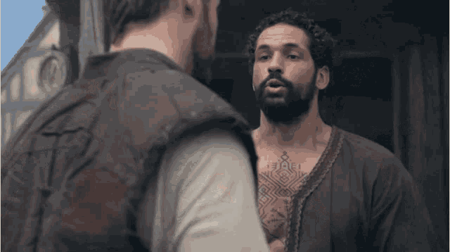 a man with a tattoo on his chest is talking to another man with a beard