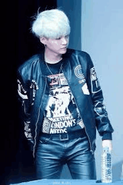 a man with white hair is standing in front of a table wearing a leather jacket and holding a bottle of water .