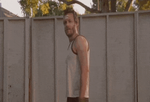 a man in a tank top stands in front of a fence