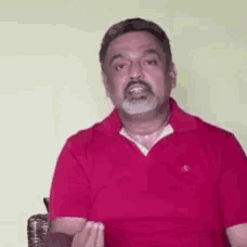 a man with a beard is sitting in a chair wearing a red shirt and making a funny face .