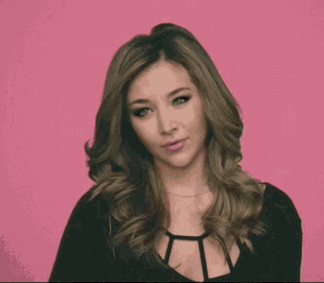 a woman with long hair is wearing a black top and making a funny face against a pink background .