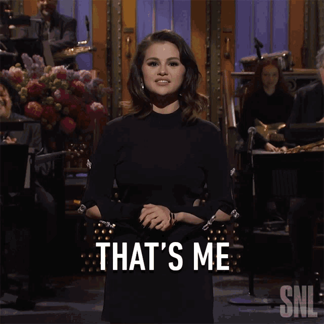 a woman in a black dress stands in front of a snl band and says that 's me