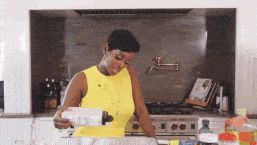 a woman in a yellow tank top is pouring something into a pan in a kitchen
