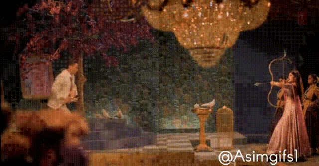 a woman in a pink dress is holding a bow and arrow in front of a chandelier