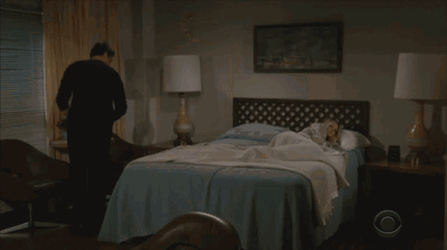 a woman stands next to a man laying on a bed that has a cbs logo on it