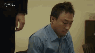 a man in a blue shirt is sitting in a chair while a man in a suit stands behind him .