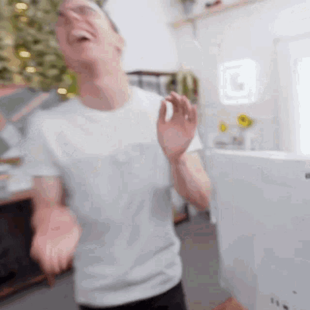 a man in a white t-shirt is standing in front of a computer monitor and laughing .
