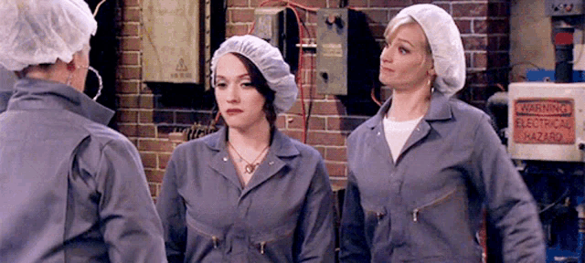three women in overalls are standing next to each other in front of a warning sign .