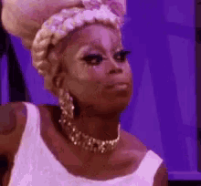 a drag queen wearing a white dress and a necklace is making a face .