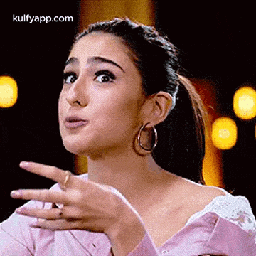 a woman in a pink shirt and hoop earrings is making a funny face with her hand .