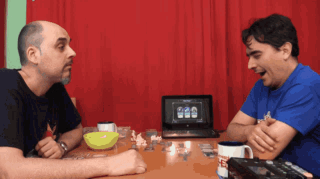 two men are sitting at a table with a laptop and a bowl of popcorn
