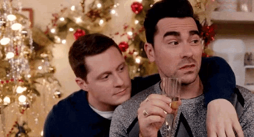 two men are sitting in front of a christmas tree one is holding a glass of champagne