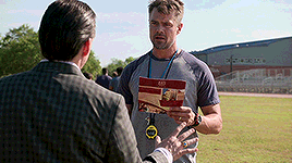 a man in a suit is talking to another man in a field while holding a booklet .