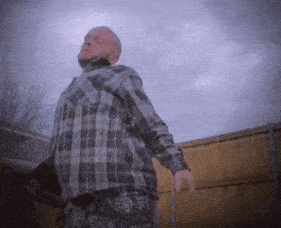 a man in a plaid shirt stands in front of a wooden fence