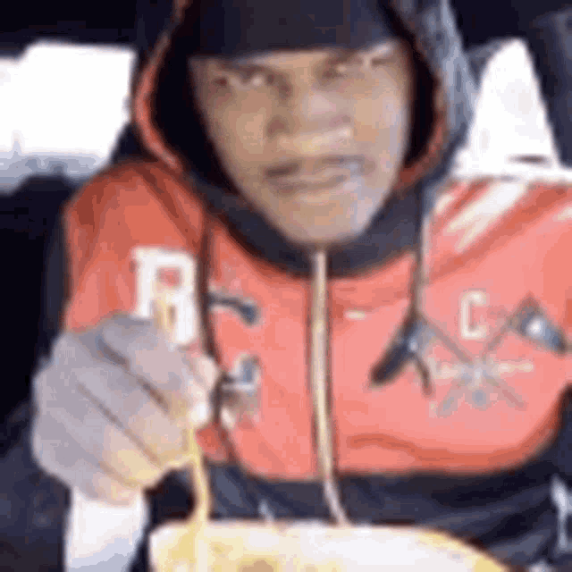 a man in a red hoodie is eating noodles with a fork in a car .
