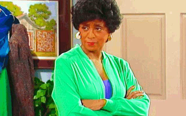 a woman in a green sweater is standing with her arms crossed and looking at the camera .