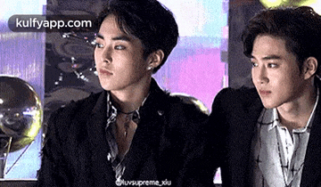 two young men in suits are sitting next to each other and looking at each other .