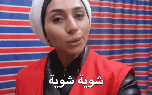 a woman wearing a red vest and a white head scarf stands in front of a striped curtain
