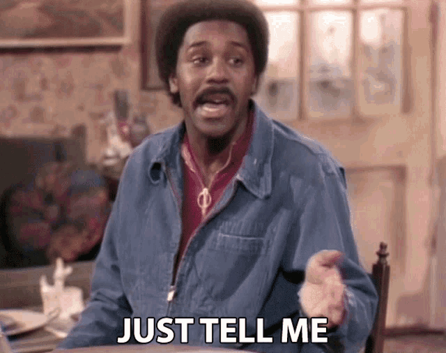 a man in a denim jacket is sitting at a table and saying just tell me