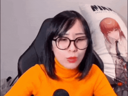 a woman wearing glasses and an orange turtleneck is sitting in a chair with a microphone .