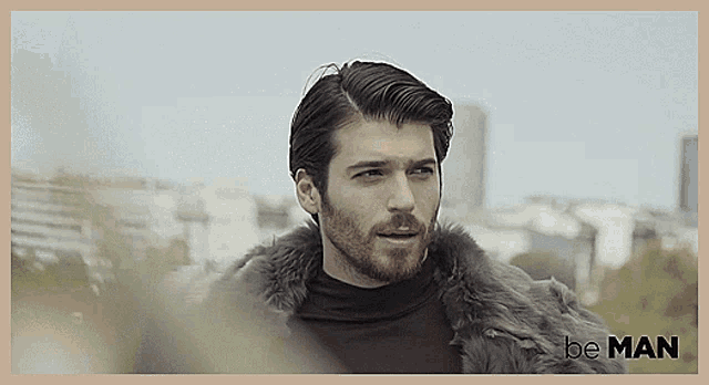 a man with a beard wearing a fur coat and a be man logo