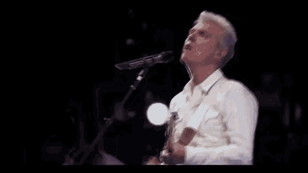 a man in a white shirt is singing into a microphone while playing a guitar .