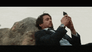 a man in a suit and tie is sitting on a rock taking a picture of himself with his cell phone .