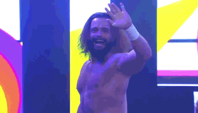 a shirtless wrestler with long hair and a beard waving his hand