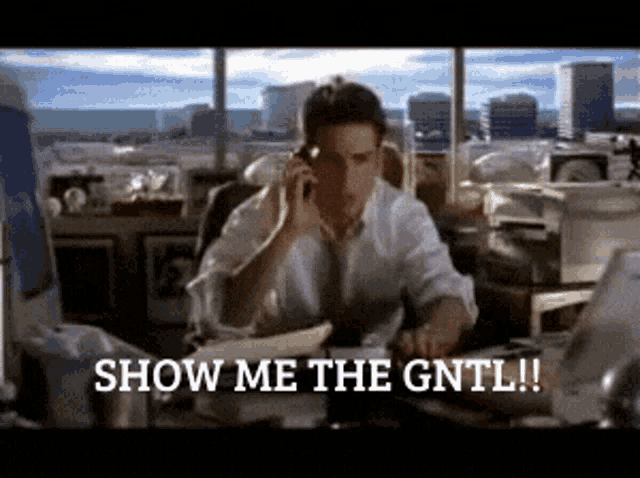 a man sitting at a desk talking on a cell phone with the words show me the gntl written below him