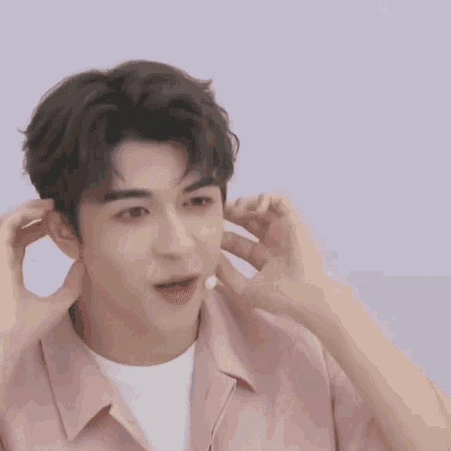 a young man in a pink shirt is covering his ears with his hands while talking into a microphone .