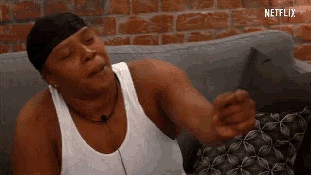 a woman in a white tank top is sitting on a couch with her eyes closed and pointing .