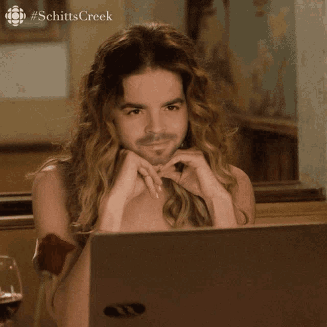 a man with long hair is sitting in front of a laptop with schittscreek written on the bottom