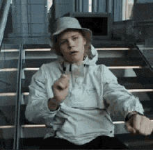 a young man is sitting on a set of stairs wearing a hat and a jacket .