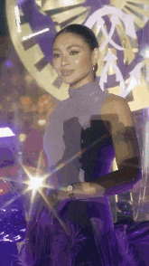 a woman in a purple dress is standing in front of a clock and looking at the camera .
