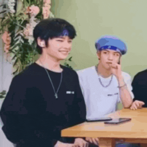two young men are sitting at a table with flowers in the background . one of the men is wearing a blue beret .