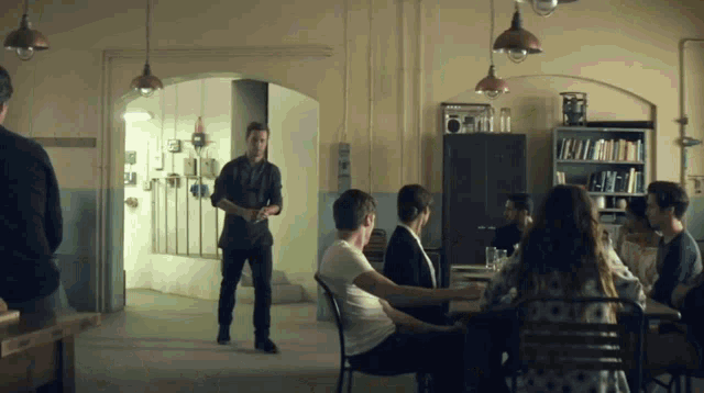 a group of people are sitting around a table with a man standing in the middle