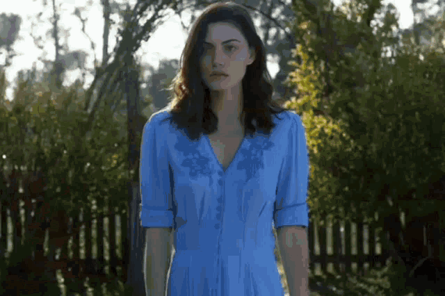 a woman in a blue dress is standing in front of a fence in a park .