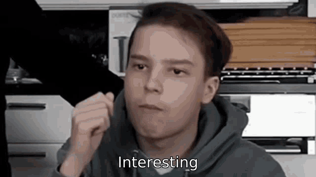 a young man in a blue hoodie is making a funny face in a kitchen and the words `` interesting '' are visible .