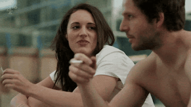 a man and a woman are sitting next to each other . the woman is smoking a cigarette and the man is shirtless .