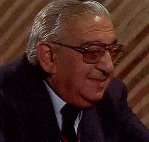 an older man wearing glasses and a suit holds his hand to his mouth